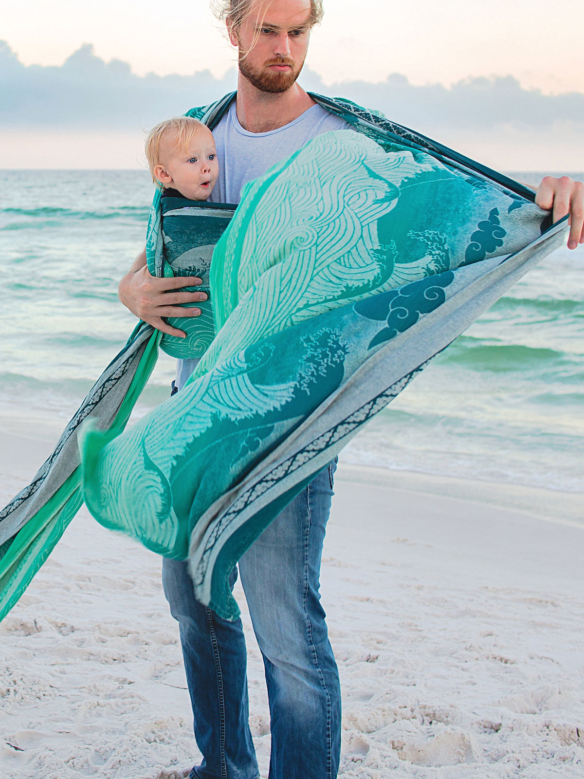 Man holding a baby wrapped in a teal woven wrap on a beach.