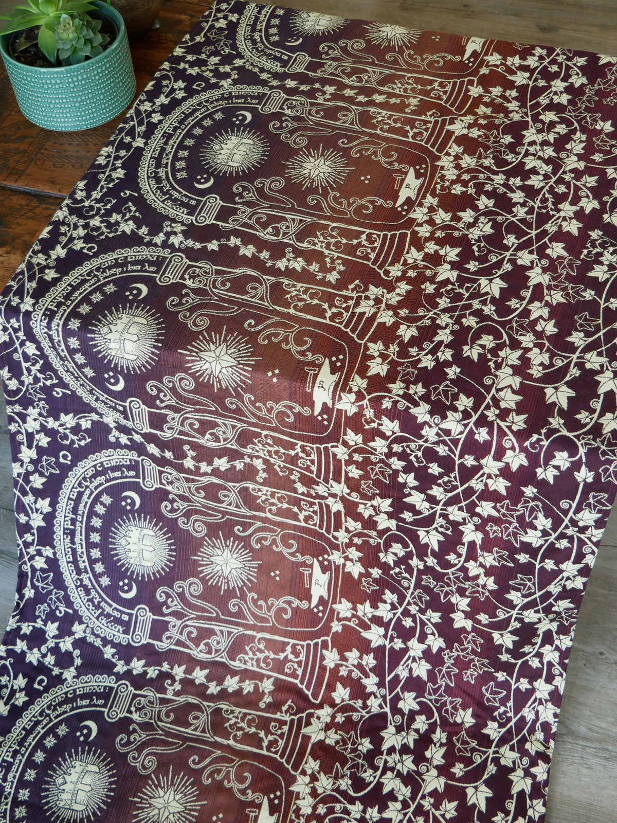 Decorative patterned fabric in dark autumnal tones on a wooden surface with a plant in the background.