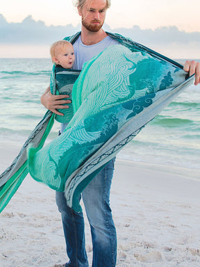 Man holding a baby wrapped in a teal woven wrap on a beach.