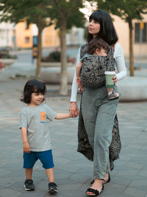 Woman walking with a child in Prowl Savannah Baby Wrap and another child holding hands on a sidewalk.